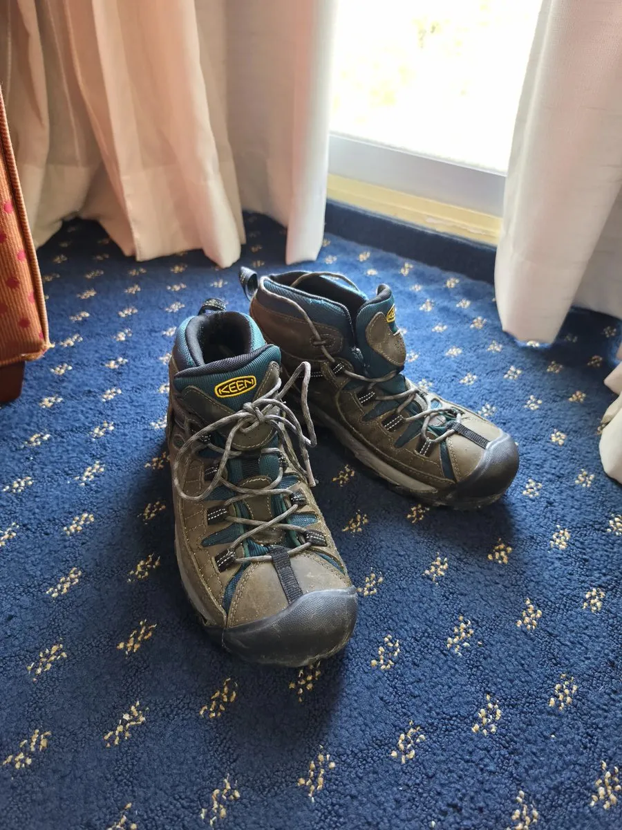 A worn pair of Keen hiking boots with teal and tan panels, laces loose, sitting on a blue carpet by a window