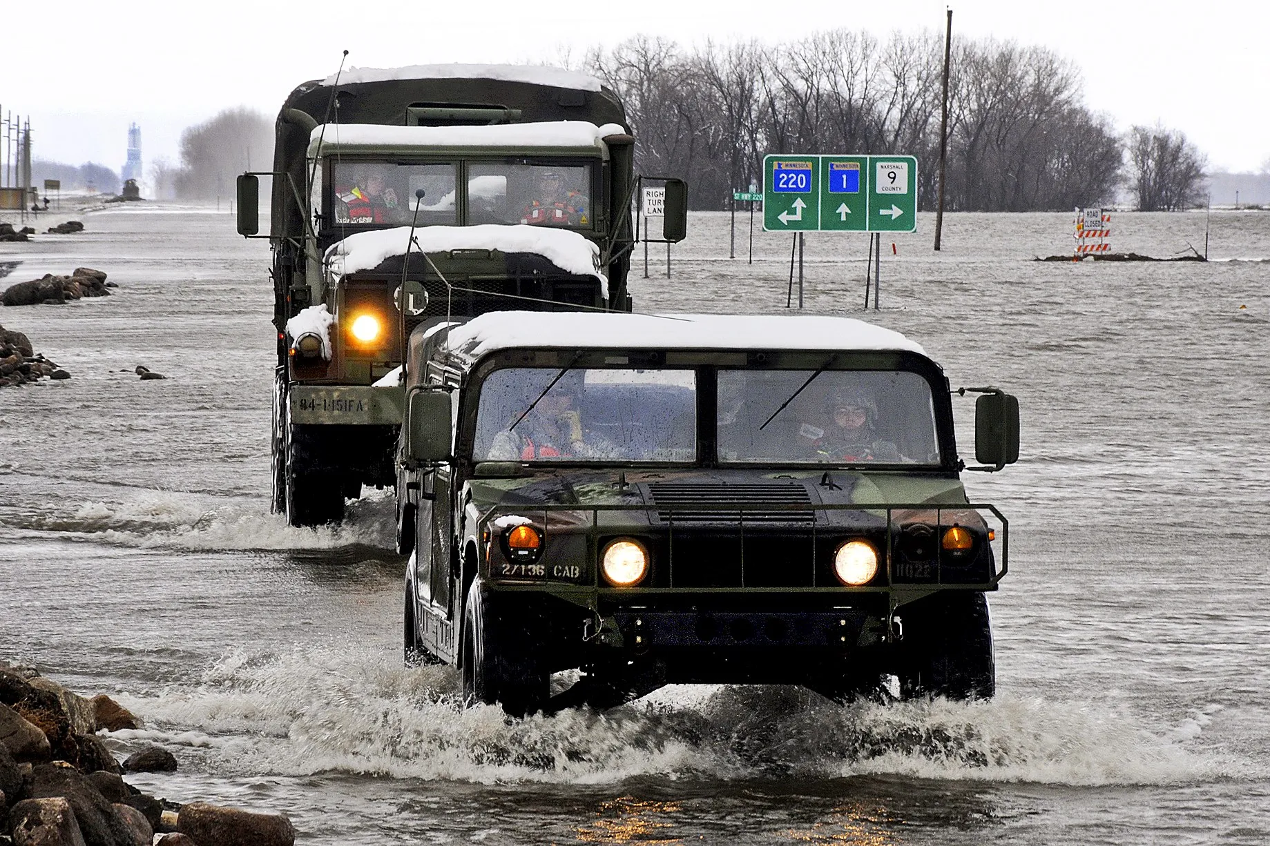Two snow-topped Minnesota National Guard trucks drive through floodwater covering a highway near Oslo, with road signs standing in the water behind them