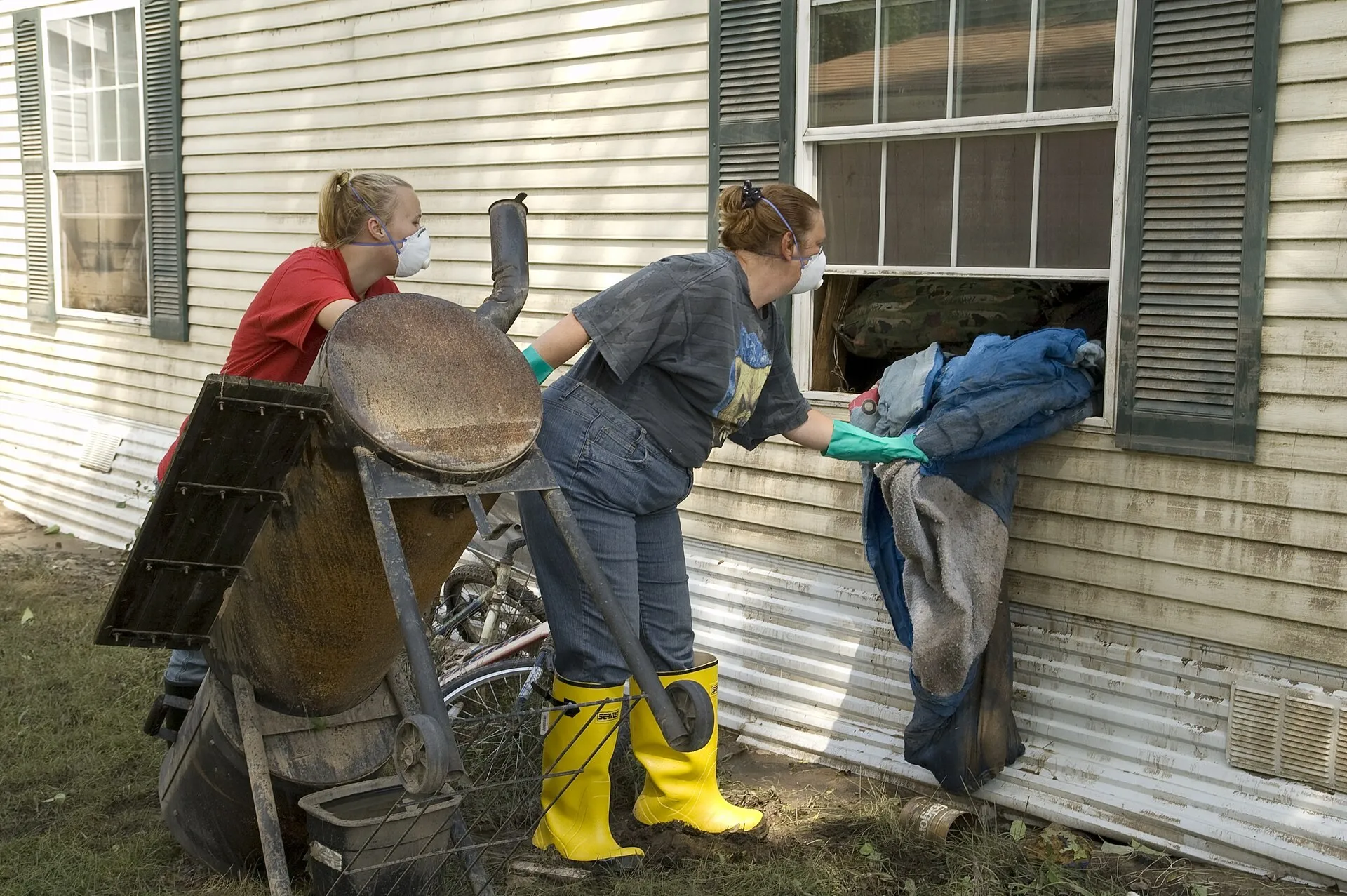 Two residents in masks and rubber boots pass belongings out through the window of a flooded house in Rushford