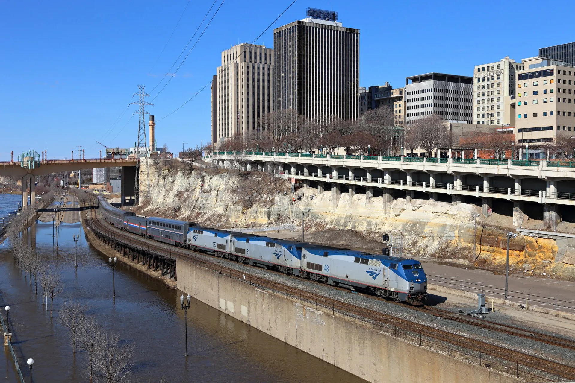 An Amtrak train passes below downtown St. Paul on a raised track, with Shepard Road and its streetlights under brown floodwater beside it